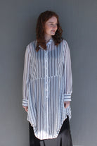A woman with curly brown hair wearing a white button-up shirt with thin black vertical stripes and a high-low hem, paired with black bottoms, standing against a plain gray wall.