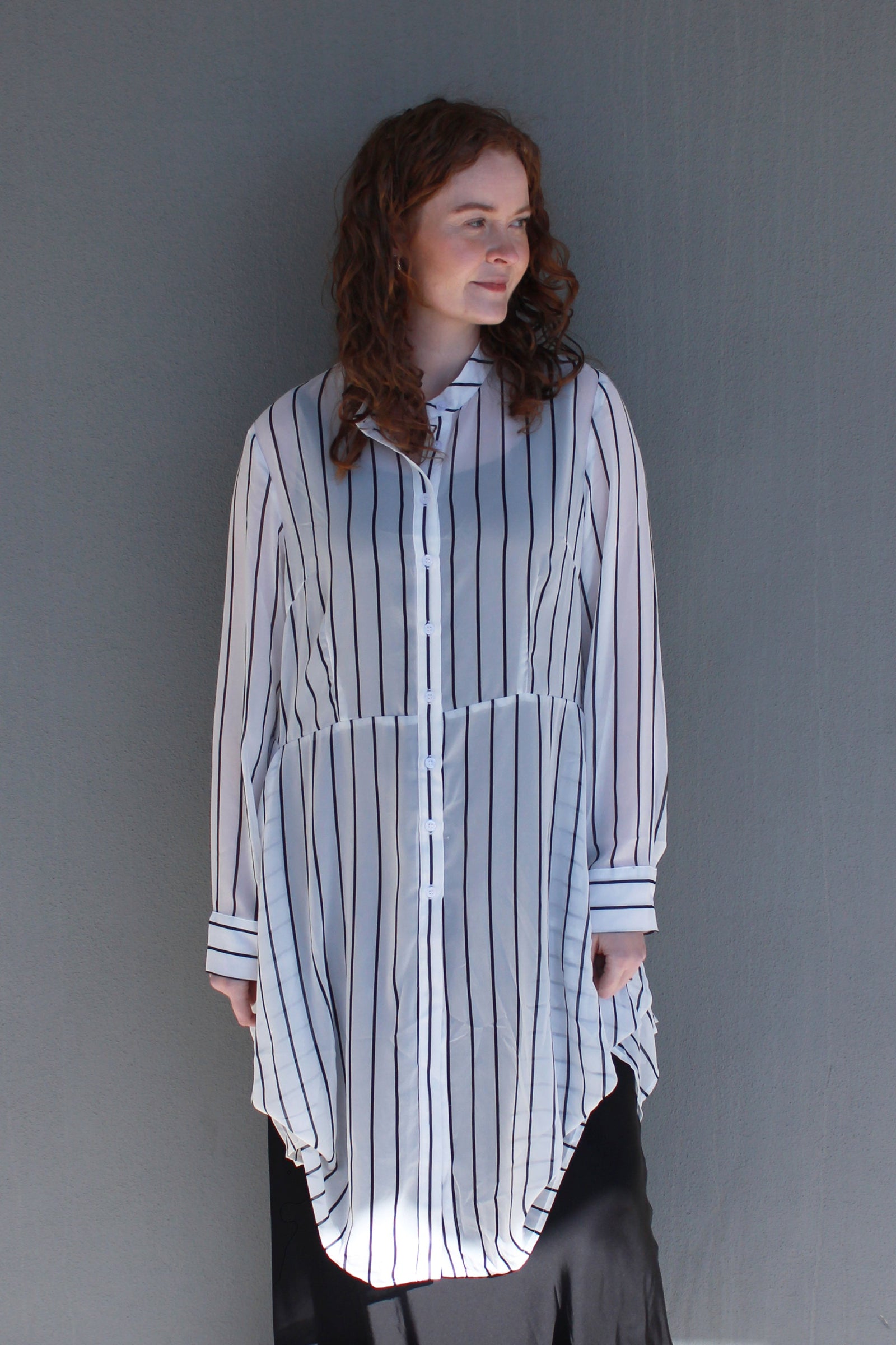 A woman with curly brown hair wearing a white button-up shirt with thin black vertical stripes and a high-low hem, paired with black bottoms, standing against a plain gray wall.