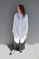 A woman stands against a plain gray wall wearing a loose white button-front shirt with thin vertical navy stripes and a long dark skirt. She is photographed in bright sunlight with a hard shadow on the ground.