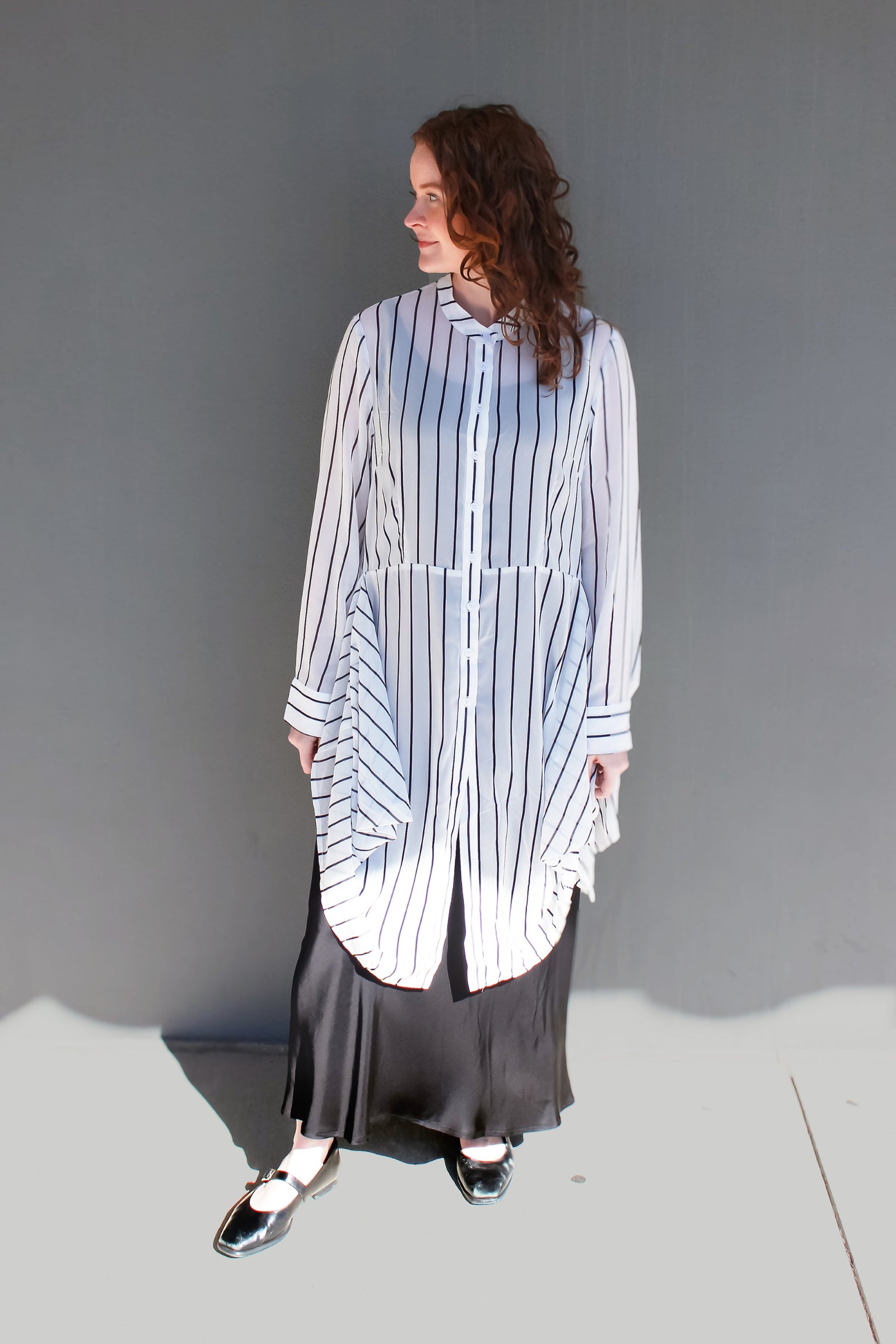 A woman stands against a plain gray wall wearing a loose white button-front shirt with thin vertical navy stripes and a long dark skirt. She is photographed in bright sunlight with a hard shadow on the ground.