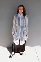 A woman modeling a long-sleeve button-front shirt dress with vertical blue and white stripes, worn over a black maxi skirt and black Mary Jane shoes, standing against a gray wall.