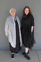 Two women standing side-by-side outdoors against a gray wall. The woman on the left has short blonde hair and wears a white button-up shirt with thin black vertical stripes over a black dress and black sneakers.
