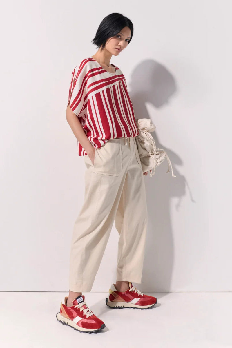 A model stands against a white studio wall wearing a red and white vertically striped short-sleeve top, cream-colored wide-leg trousers, and red sneakers, while holding a cream jacket.
