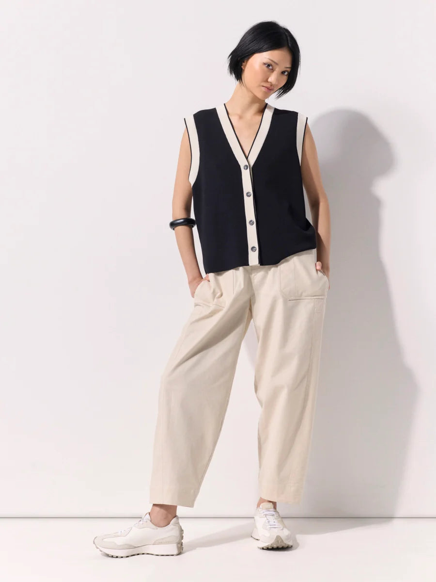 A woman modeling a sleeveless black knit vest with cream trim and buttons, paired with cream wide-leg trousers and white sneakers, standing against a plain white wall.