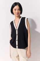 Woman wearing a black buttoned up sleeveless knit vest with white contrast trim and black buttons against a white wall.