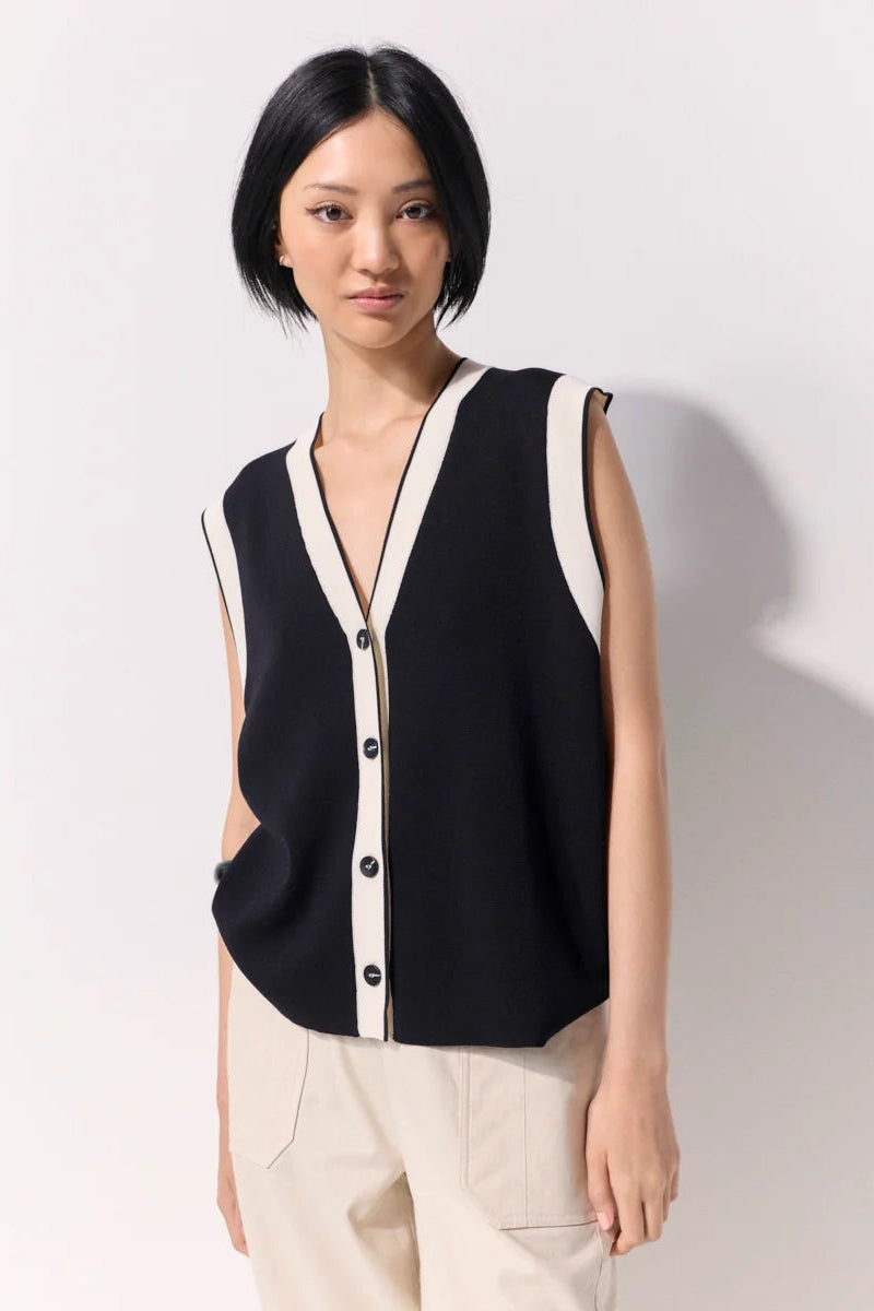 Woman wearing a black buttoned up sleeveless knit vest with white contrast trim and black buttons against a white wall.