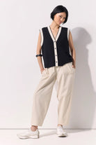 Woman wearing a black buttoned up sleeveless knit vest with white contrast trim and black buttons against a white wall.