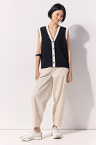 Woman wearing a black buttoned up sleeveless knit vest with white contrast trim and black buttons against a white wall.