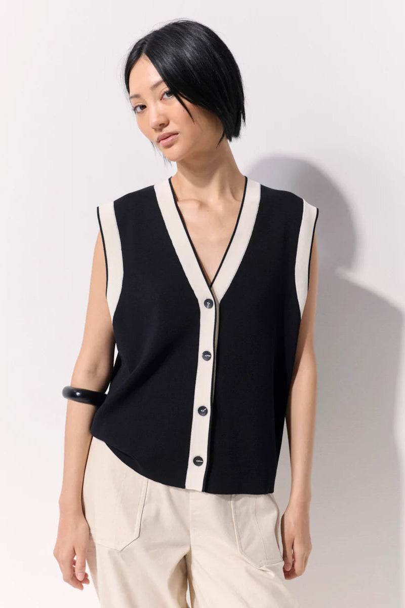 Woman wearing a black buttoned up sleeveless knit vest with white contrast trim and black buttons against a white wall.