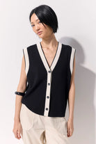 Woman wearing a black buttoned up sleeveless knit vest with white contrast trim and black buttons against a white wall.