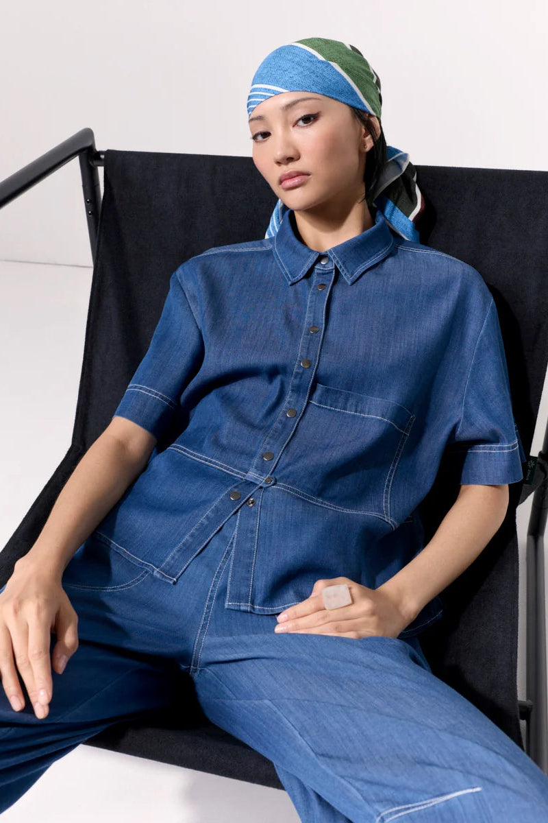 Woman wearing a short sleeve light denim shirt with collar and front left side pocket with contrast stitching against a white background.