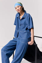 Woman wearing a short sleeve light denim shirt with collar and front left side pocket with contrast stitching against a white background.