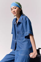 Woman wearing a short sleeve light denim shirt with collar and front left side pocket with contrast stitching against a white background.