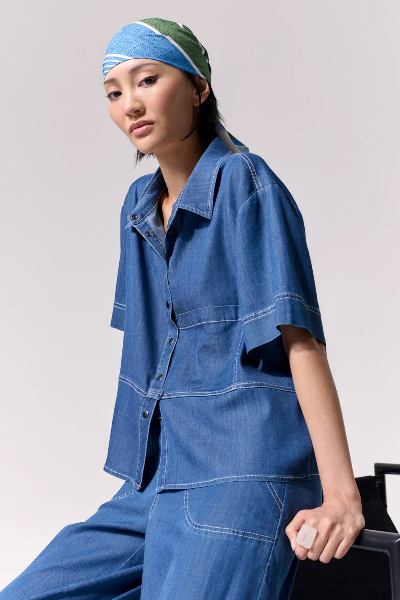 Woman wearing a short sleeve light denim shirt with collar and front left side pocket with contrast stitching against a white background.