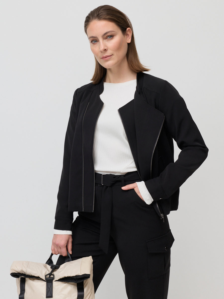 Woman wearing a black jacket and pants with a white top, holding a beige backpack on a light gray background.