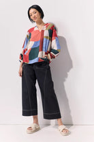 A model wearing a colorful geometric print blouse with wide three-quarter sleeves, black wide-leg cropped pants, and beige slide sandals, standing against a plain white wall.