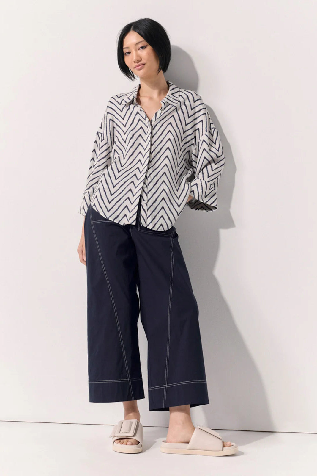 A woman stands against a plain light wall wearing a loose black-and-white zigzag patterned button-up shirt, black wide-leg cropped pants with contrast stitching, and light beige slide sandals.