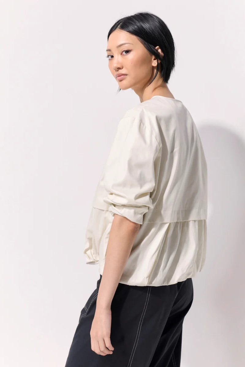 Woman wearing a cropped jacket with pushed up sleeves and no collar against a white background.