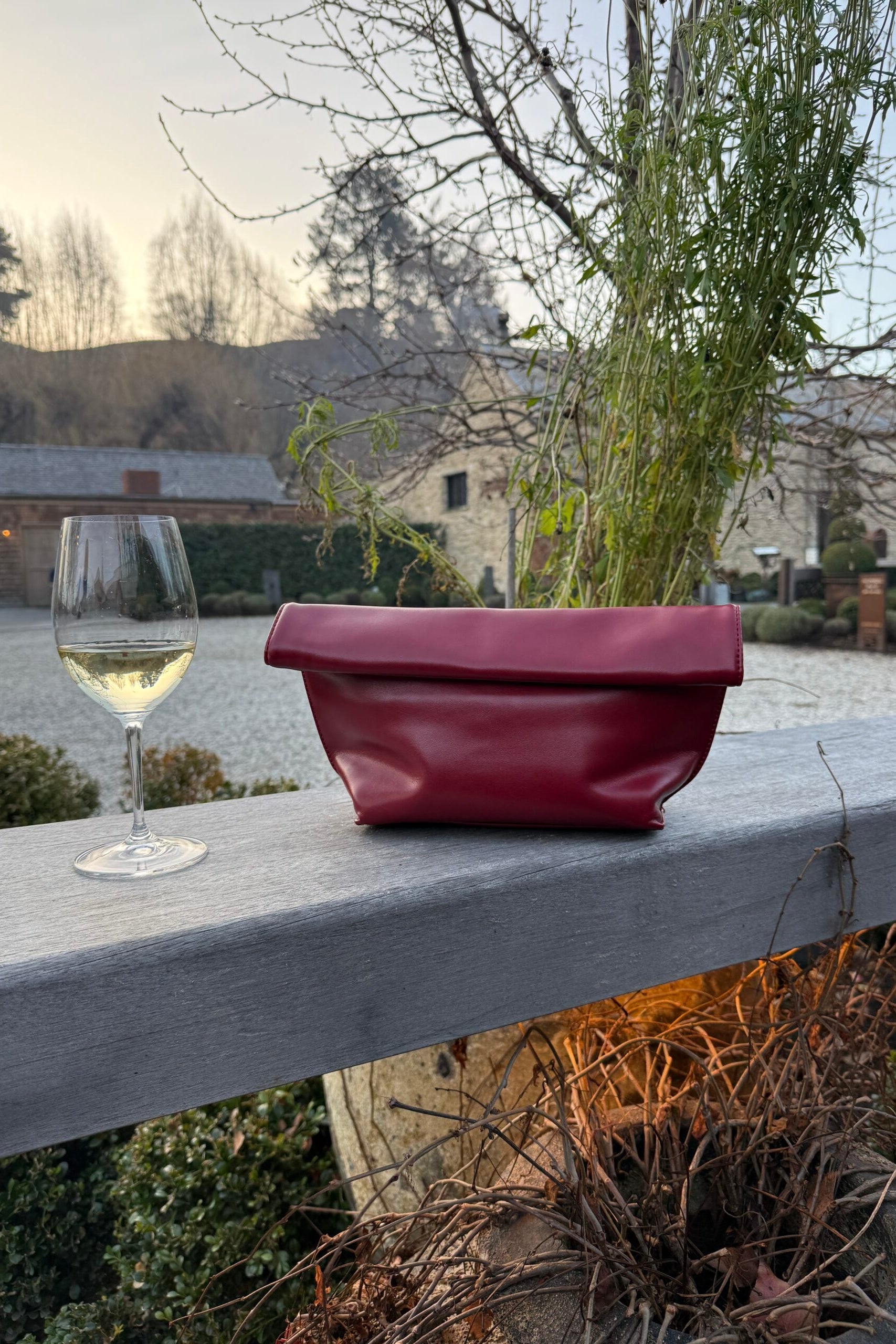 A burgundy leather clutch bag sits on a gray wooden railing next to a glass of white wine, with an outdoor garden and stone building in the background.