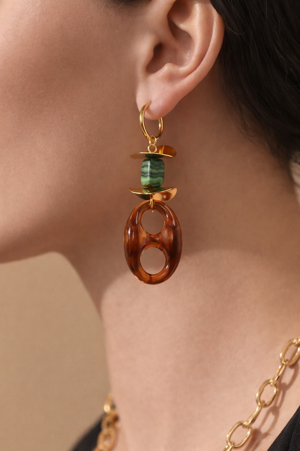 A close-up of a model's ear wearing a gold hoop earring with a green marbled bead and a large oval amber-brown resin drop.