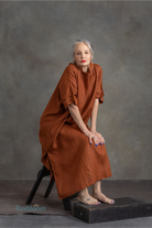 Woman in a rust-colored kaftan sitting on a black stool against a gray background