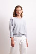 Woman wearing a light gray sweater and white pants against a plain background