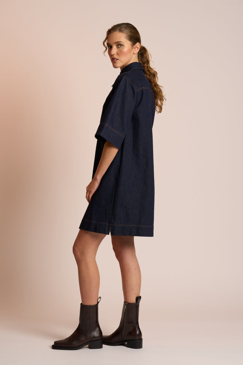 Woman wearing a navy dress and black boots against a beige background side angle