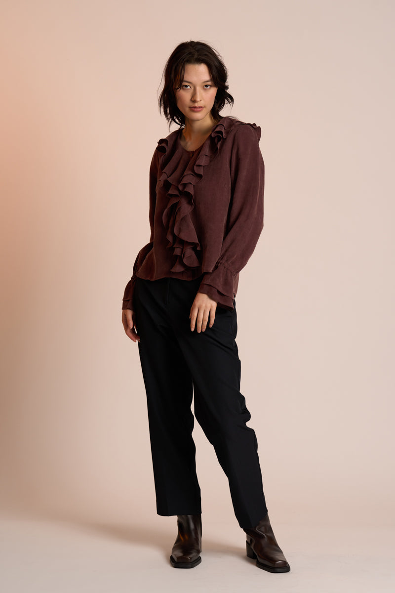 Woman wearing a brown ruffled blouse and black pants against a beige background
