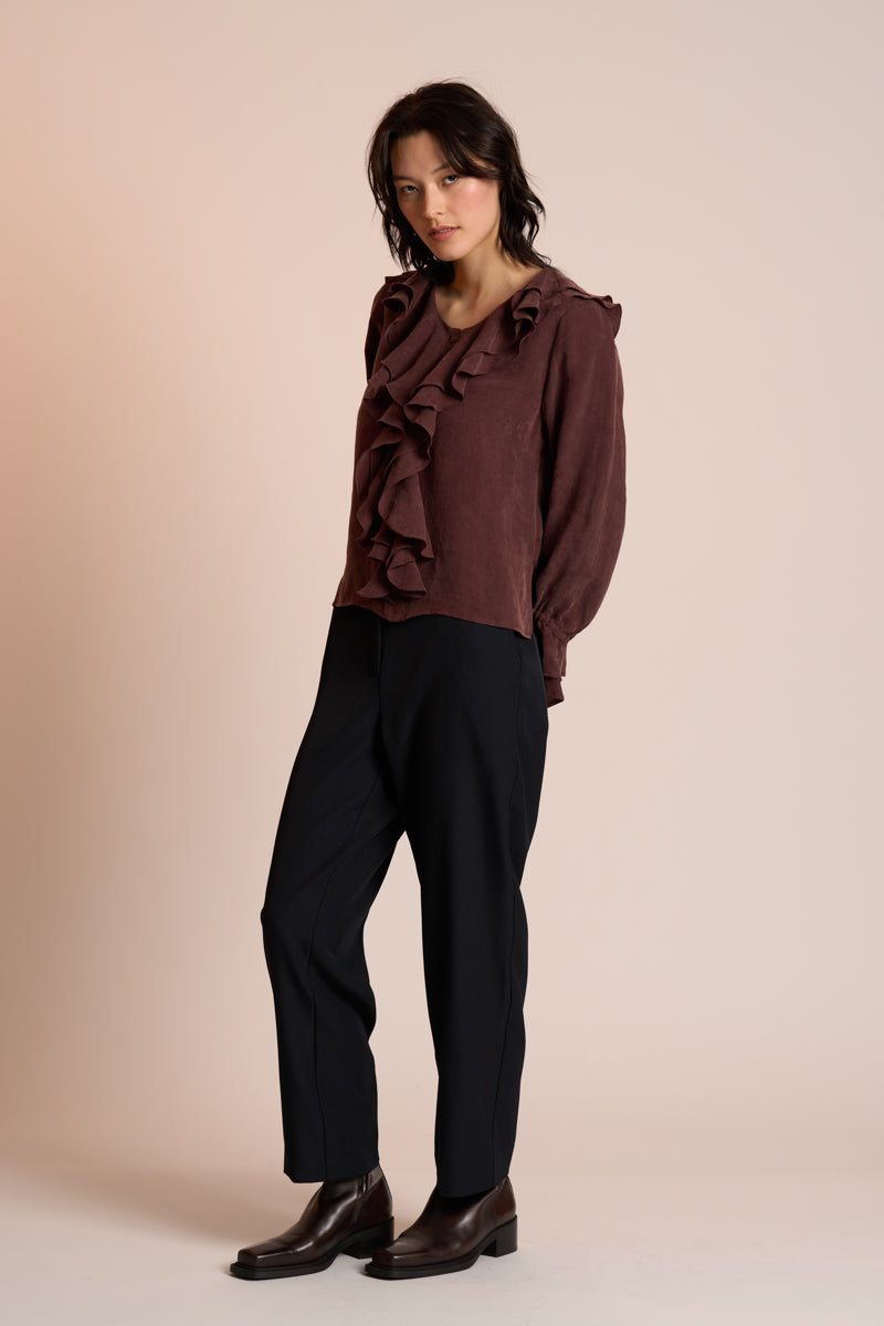 Woman wearing a brown ruffled blouse and black pants against a beige background