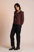 Woman wearing a brown ruffled blouse and black pants against a beige background