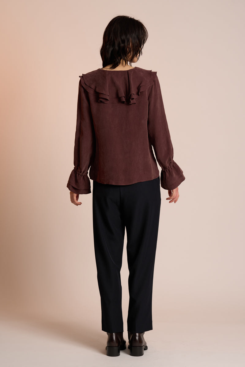 Person wearing a brown blouse with ruffled details and black pants on a beige background back view