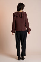 Person wearing a brown blouse with ruffled details and black pants on a beige background back view