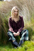 Woman sitting in tall grass wearing a purple blouse and plaid pants.