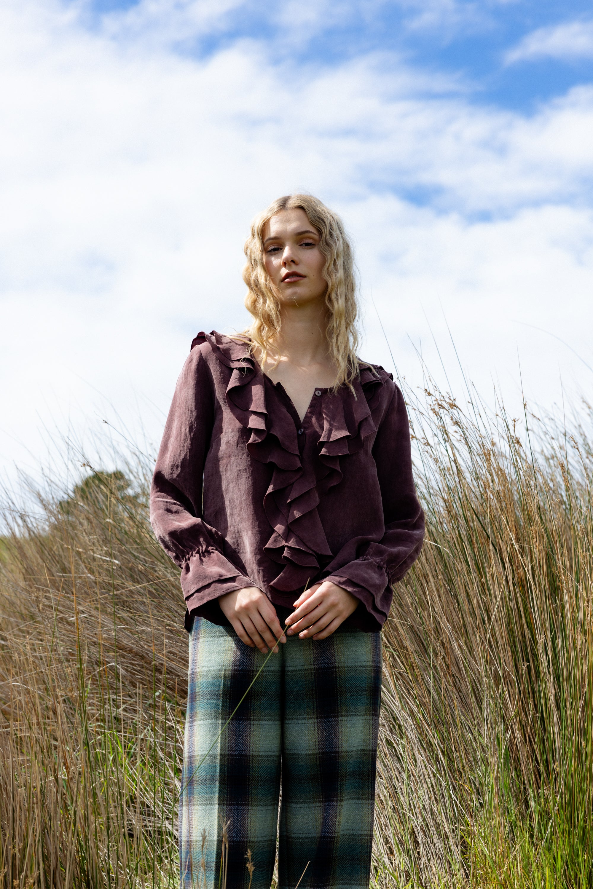 Woman wearing a ruffled blouse and plaid pants standing in tall grass with a blue sky.