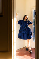 Woman in a blue dress standing in a sunlit room with a door open.