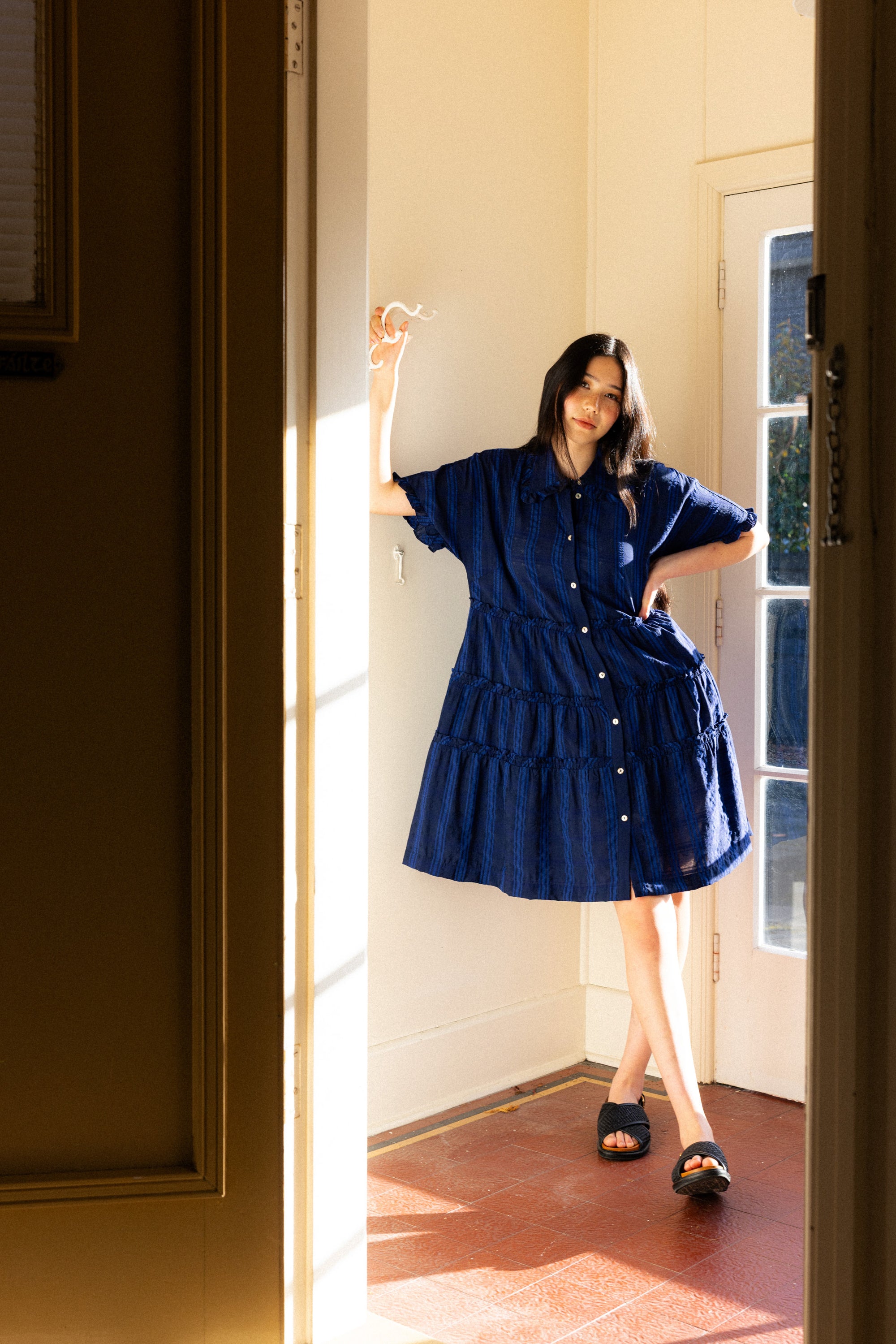 Woman in a blue dress standing in a sunlit room with a door open.