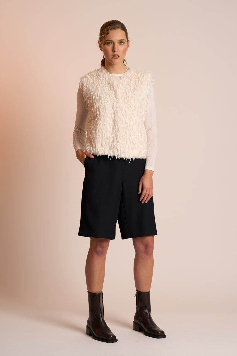 Woman wearing an off white gilet, black shorts, and brown boots on a beige background