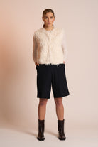 Woman wearing an off white gilet, black shorts, and brown boots on a beige background