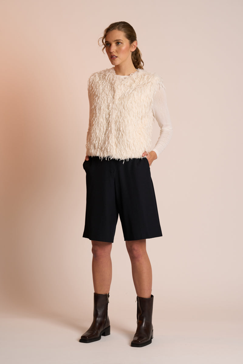Woman wearing an off white gilet, black shorts, and boots on a beige background