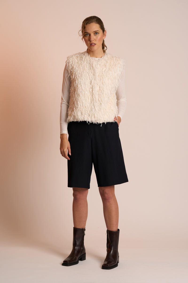 Woman wearing an off white gilet, black shorts, and brown boots on a beige background