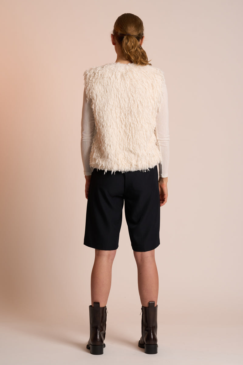 Woman wearing an off white gilet, black shorts, and brown boots on a beige background back angle