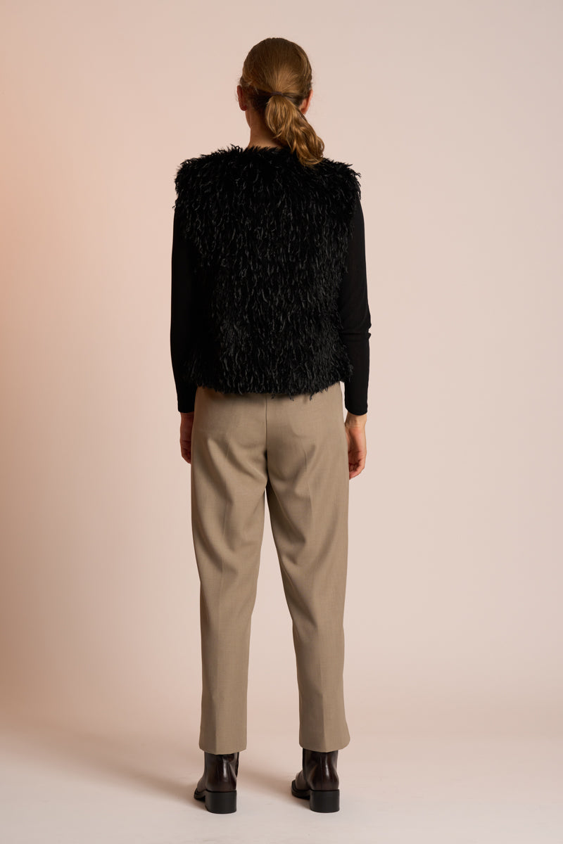 Woman wearing a black gilet over a black top with beige pants on a beige background back angle