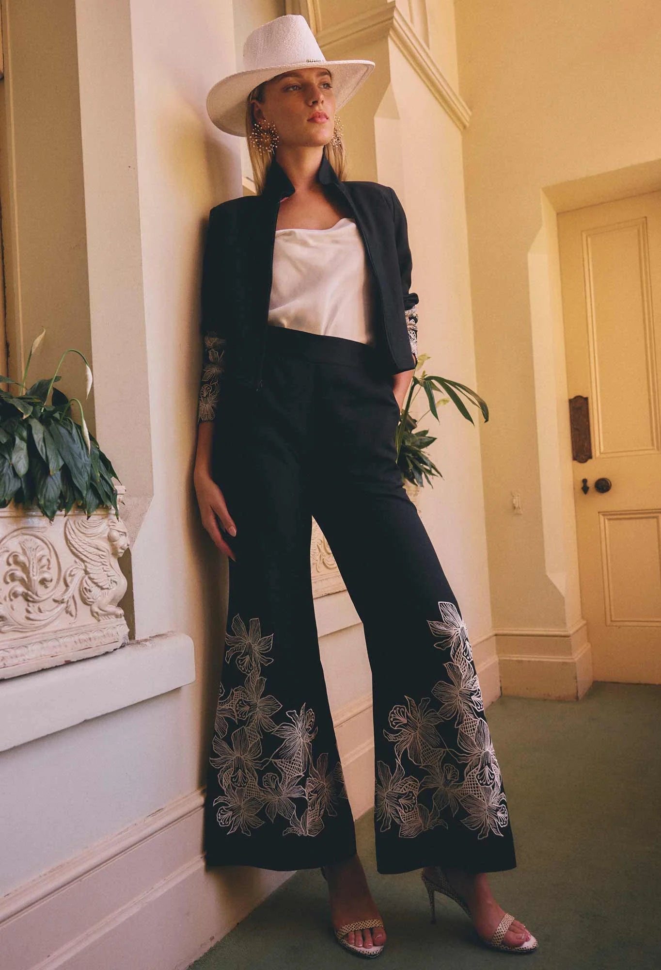 women wearing a pair of wide leg black pants with floral embroidery and a matching black jacket with a white hat 