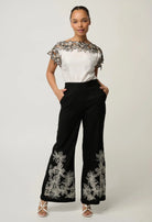 women wearing a white top with black floral embroidery and black pants with with white embroidery on a plain background