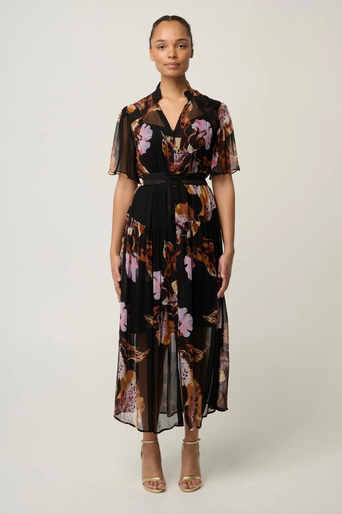 Woman wearing a black floral floaty chiffon dress with tie waist and short sleeves and gold sandals against a white background
