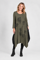 A woman modeling a long-sleeve olive green tunic dress with black abstract graphic prints on the front, styled with black leggings and black ankle boots against a light gray studio background.