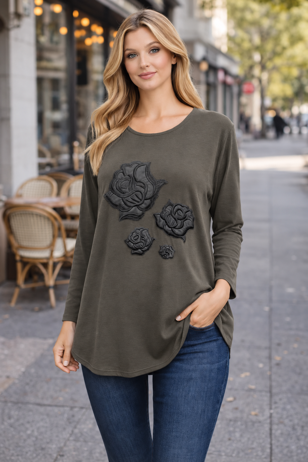 A woman modeling an olive green long-sleeve tunic top with black rose appliques, paired with blue jeans, standing on a city sidewalk.