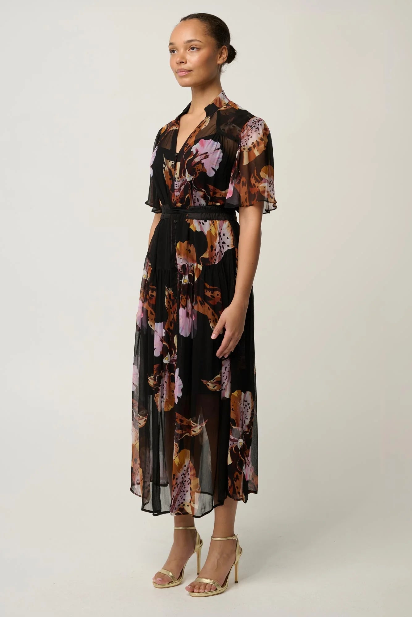 Woman wearing a black floral floaty chiffon dress with tie waist and short sleeves and gold sandals against a white background