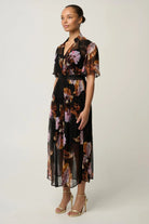 Woman wearing a black floral floaty chiffon dress with tie waist and short sleeves and gold sandals against a white background