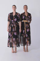 Woman wearing a black floral floaty chiffon dress with tie waist and short sleeves and gold sandals against a white background